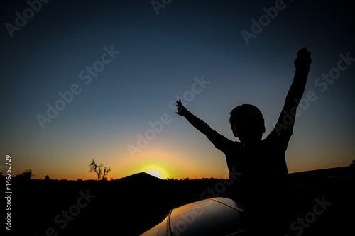 Silhouette of child with arms up on top of a vehicle in front of Pyramid Hill in Victoria, Australia at sunset
