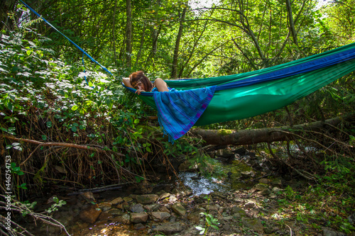 A girl lies resting in a green hammock stretched out on blue slings over a small mountain river in the shade of a dense forest. Hot summer day. Nearby there is a fallen larch and stones. General shot.