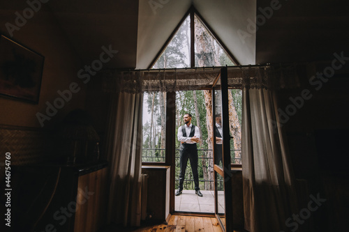 A handsome man, a bearded businessman stands in the interior on the veranda, terrace, house with a roof in the window, against the background of nature, forest.