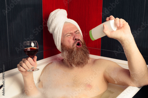 Cheerful bearded man taking bubble bath holding glass of red wine and singing into shampoo bottle with facial expression. Relaxation at home after hard day. Happy guy enjoying life.