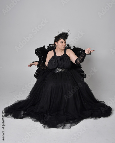 Wallpaper Mural Full length portrait of young  plus sized woman with short  hair,  wearing long black tulle gothic gown with crown and gloves, sitting pose on a ornate throne chair on light studio background. Torontodigital.ca