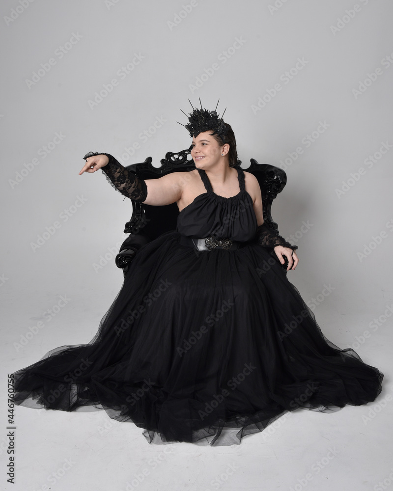Fototapeta premium Full length portrait of young plus sized woman with short hair, wearing long black tulle gothic gown with crown and gloves, sitting pose on a ornate throne chair on light studio background.