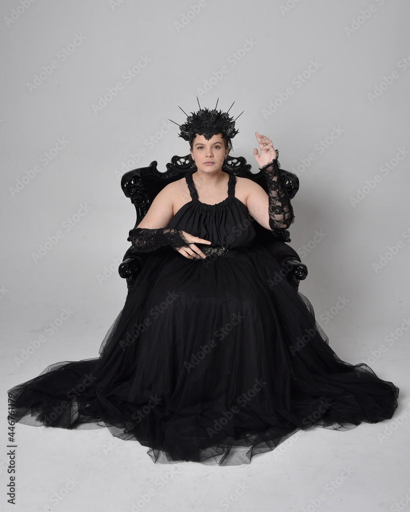 Full length portrait of young  plus sized woman with short  hair,  wearing long black tulle gothic gown with crown and gloves, sitting pose on a ornate throne chair on light studio background.