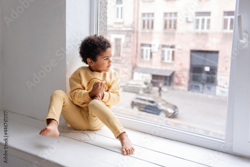 A little African American boy in yellow pajamas sits barefoot on a wide windowsill by a large white window. The child is having fun  laughing and fooling around. Happy life style childhood at home