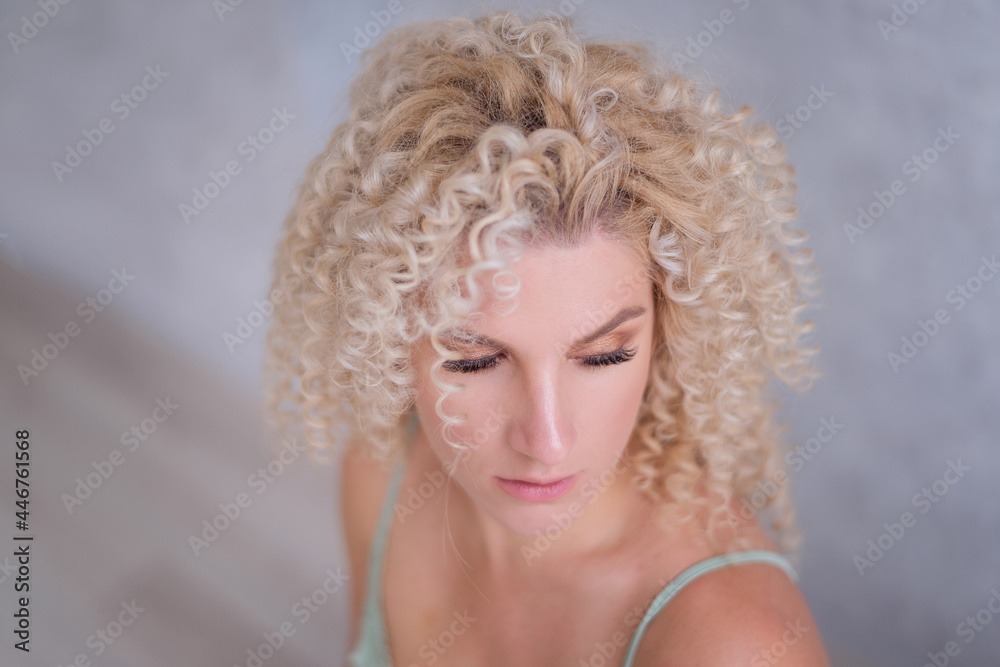 Close-up beauty portrait of caucasian young blonde woman with afro curls hairstyle with blue eyes. Girl in mint elegant dress on gray textured isolated background. Natural makeup, clear skin