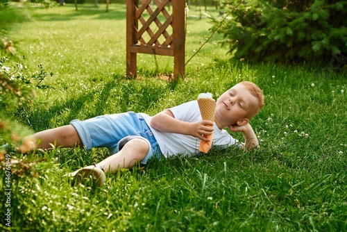 A small red-haired five-year-old boy lies on the grass in the park, eats cream ice cream in a waffle cone and rests in the shade