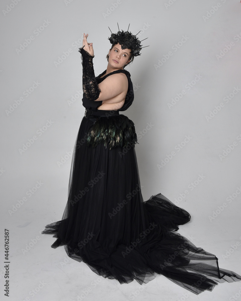 Full length portrait of young plus sized woman with short hair, wearing long black tulle gothic gown with crown and gloves, standing pose on light studio background. Full length portrait of young plus sized woman with short hair, wearing long black tulle gothic gown with crown and gloves, standing pose on light studio background.