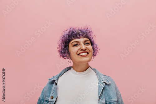 Photo of woman with stylish haircut in round big earrings  in white T-shirt and light denim jacket smiling on pink background..