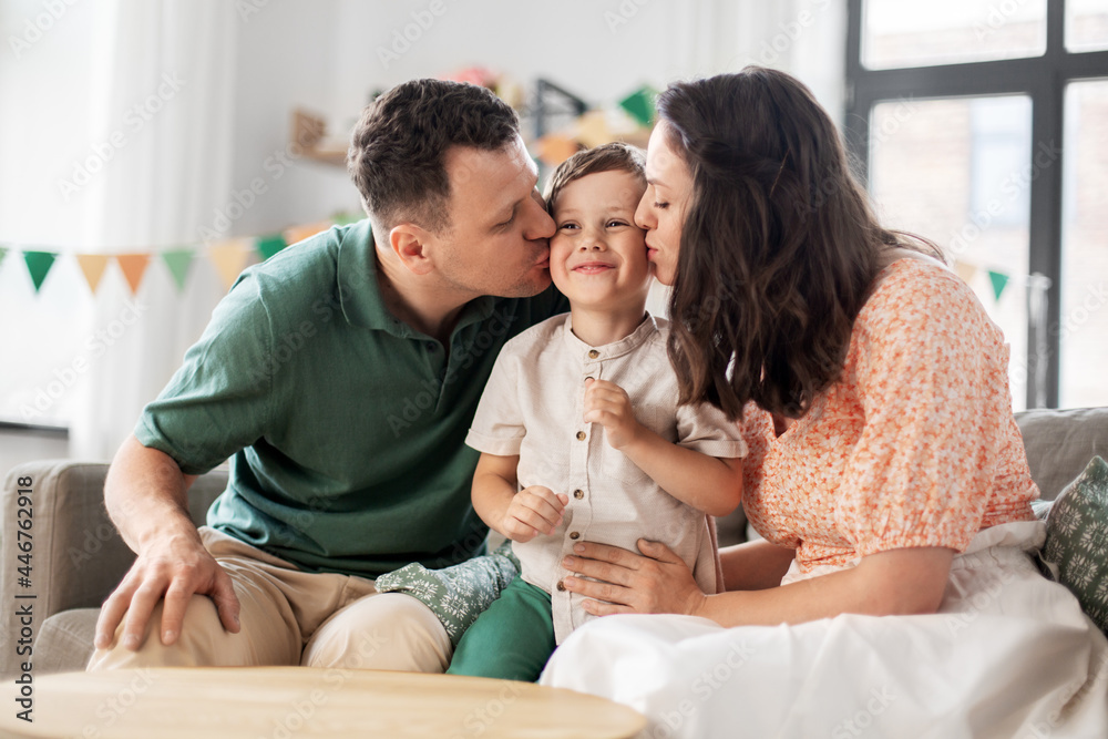family, holidays and people concept - portrait of happy mother and father kissing little son sitting on sofa at home party family, holidays and people concept - portrait of happy mother and father kissing little son sitting on sofa at home party