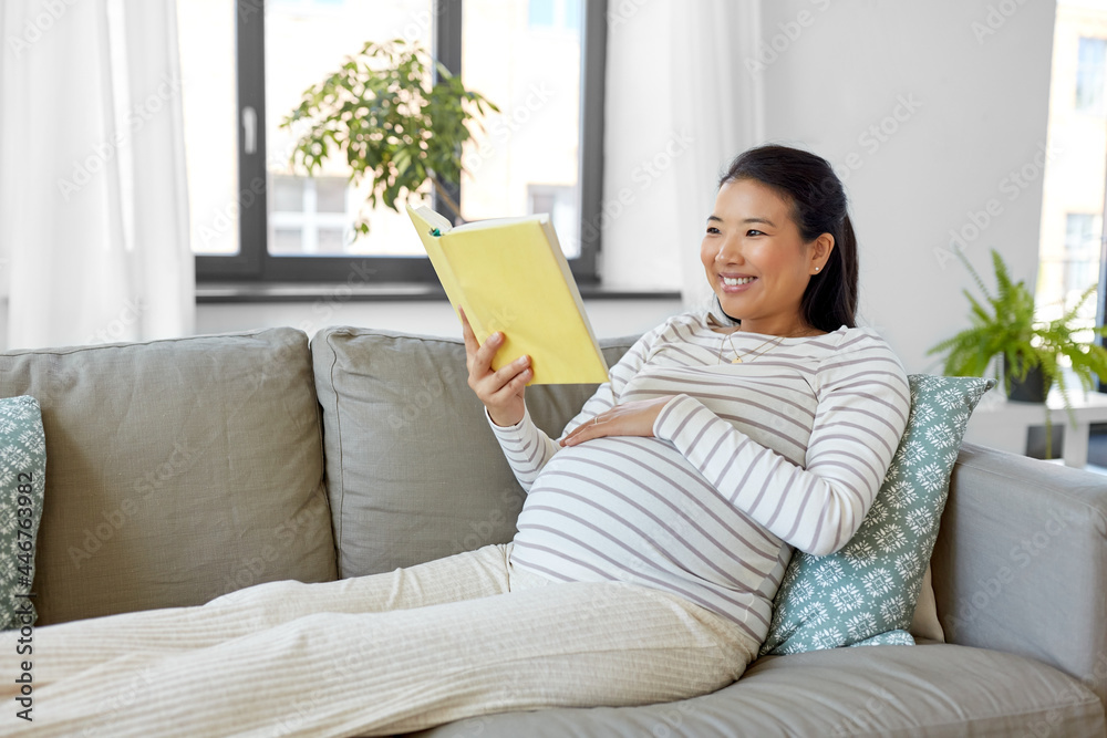 pregnancy, people and leisure concept - happy smiling pregnant asian woman sitting on sofa and reading book at home pregnancy, people and leisure concept - happy smiling pregnant asian woman sitting on sofa and reading book at home