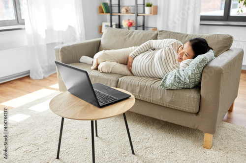 pregnancy, rest, people and expectation concept - happy smiling pregnant asian woman with laptop computer sitting on sofa at home
