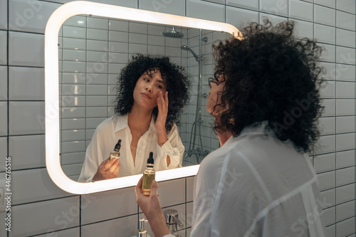 Young mixed race female gently applying face oil serum with fingers in modern bathroom. photo