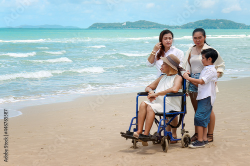 Happy disabled senior elderly woman in wheelchair spending time together with her family on tropical beach. Asian grandma, daughter and grandchildren resting and relaxing on summer holiday vacation