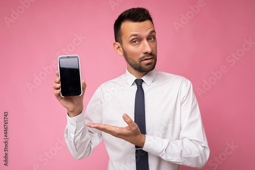 Handsome happy cool young man good looking wearing casual stylish clothes standing isolated over colourful background wall holding smartphone and showing phone with empty screen display looking at