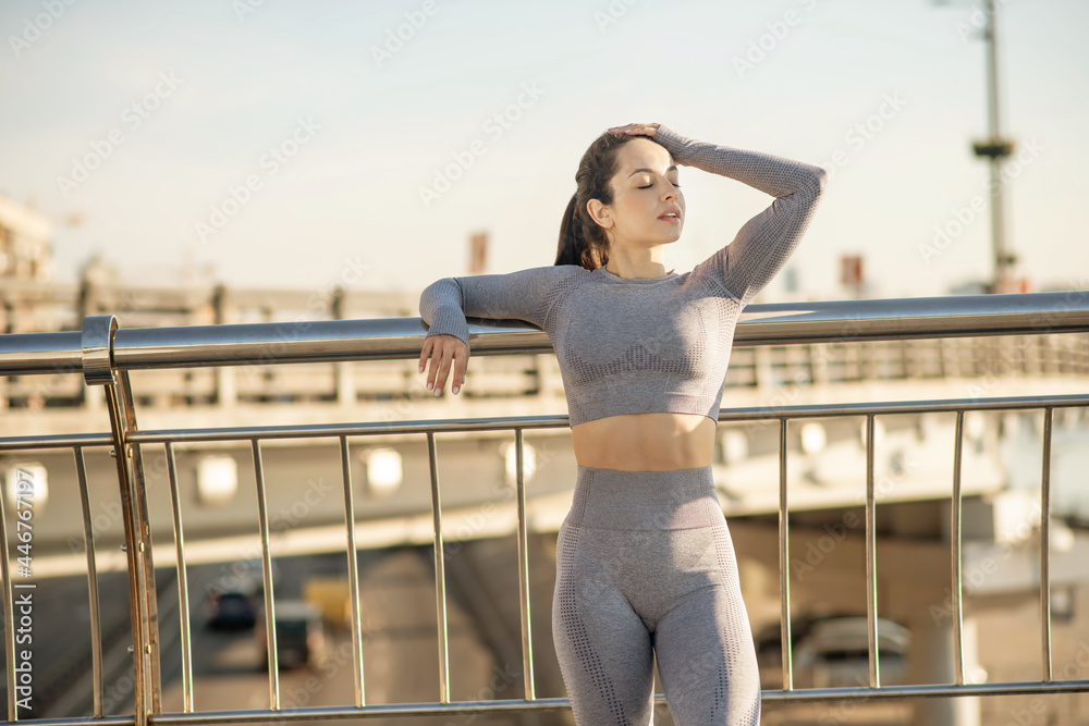 Fit girl in grey sportswear standing at the bridge and looking relaxed