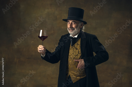 Portrait of elderly gray-haired man, gentleman, aristocrat or actor tasting wine isolated on dark vintage background. Retro style, comparison of eras concept.
