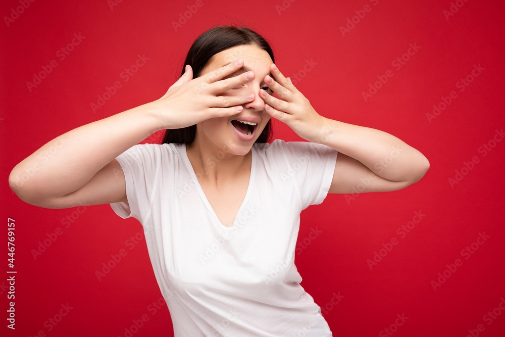 Photo of positive young beautiful brunette woman with sincere emotions wearing white t-shirt for mockup isolated over red background with empty space and covers eyes Photo of positive young beautiful brunette woman with sincere emotions wearing white t-shirt for mockup isolated over red background with empty space and covers eyes