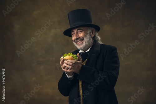 Handsome elderly gray-haired man, gentleman, aristocrat or actor eating fast food isolated on dark vintage background. Retro style, comparison of eras concept.