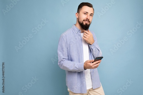 handsome young brunette unshaven man with beard wearing stylish white t-shirt and blue shirt isolated over blue background with empty space holding in hand and using phone messaging sms looking at