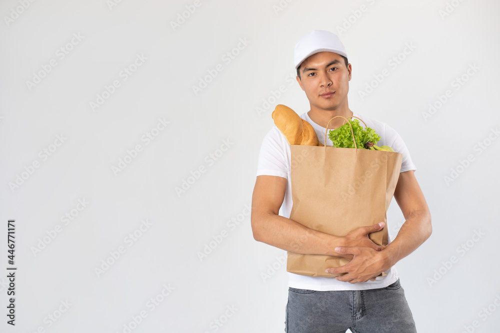 Asian delivery man in white cap holds craft bag with groceries on white background. Asian delivery man in white cap holds craft bag with groceries on white background.
