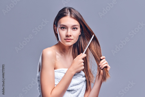 A beautiful brown-haired woman with bared shoulders looks at the camera as she brushes her hair with a wooden brush, isolated on a gray background with copy space for text. photo