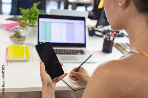 Caucasian female creative worker sitting at desk using smartphone taking notes