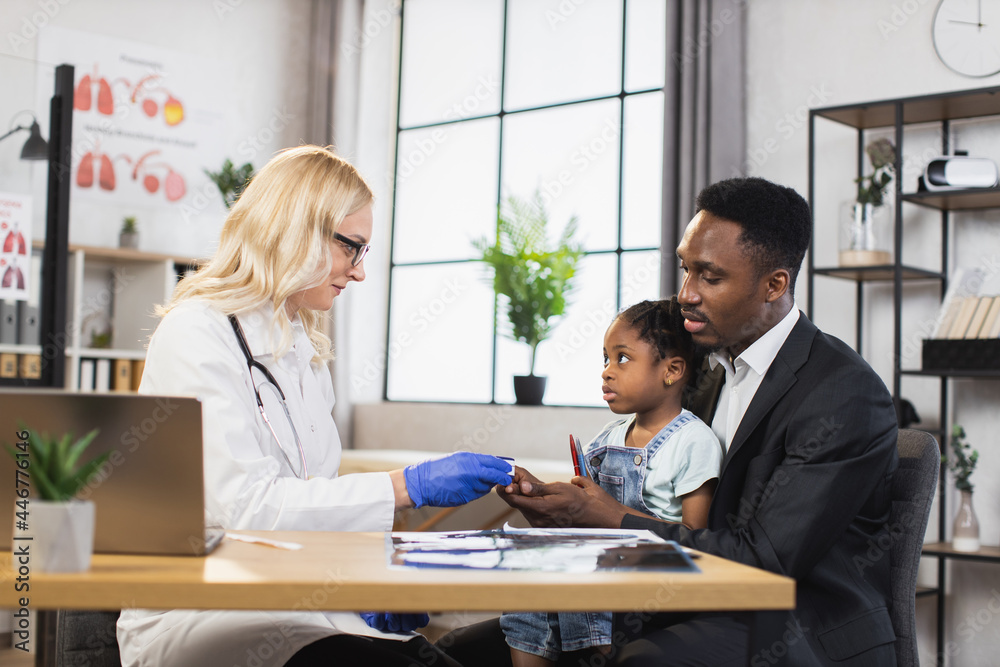Caucasian doctor checking oxygen saturation of little african girl using modern pulse oximeter. Cute patient sitting on knees af her caring father. Family visiting doctor for controlling health. Caucasian doctor checking oxygen saturation of little african girl using modern pulse oximeter. Cute patient sitting on knees af her caring father. Family visiting doctor for controlling health.