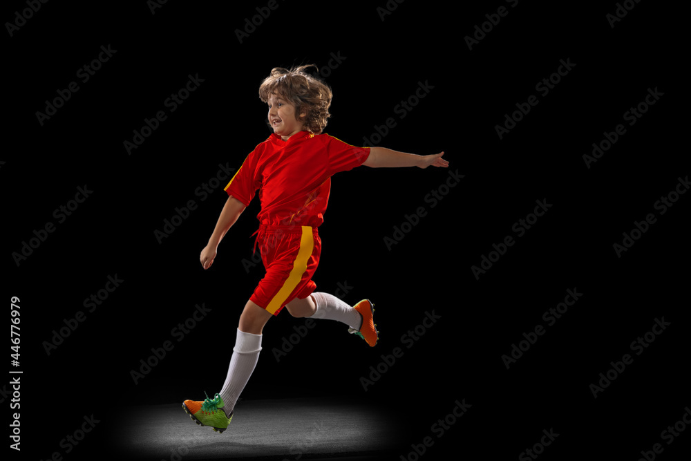 One little male football soccer player, boy training with football ball isolated on dark studio background. Concept of sport, game, hobby