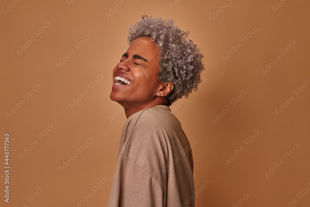Fototapeta premium Overemotive African American woman laughs loudly hears funny joke or story, being overjoyed raises palms with satisfaction, dressed in beige t shirt, amused by friend. People and emotions concept.
