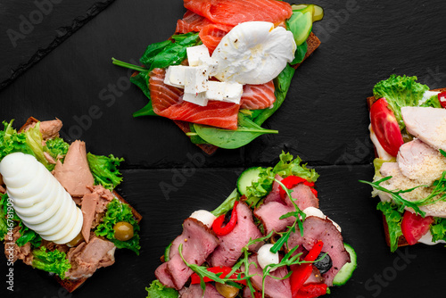 Assorted different toasts with salmon, meat, eggs, tomatoes and cheese on black stone background.Top view. Space for text. High quality photo