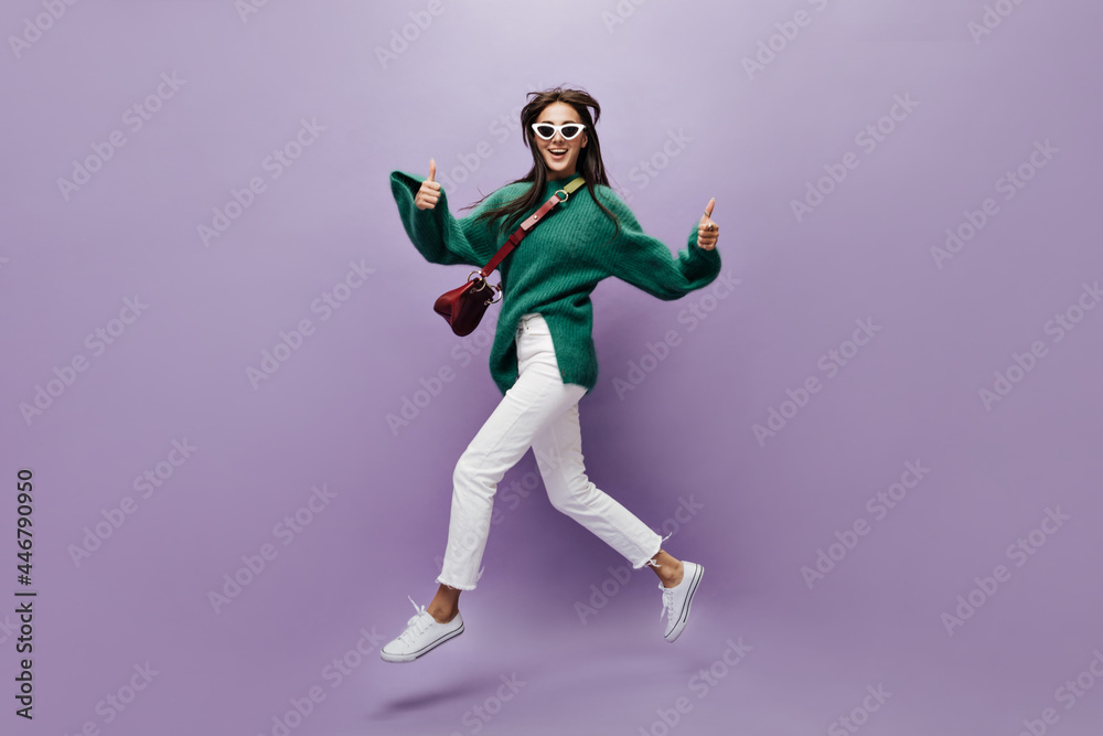 Attractive woman in stylish outfit moves and shows thumbs up. Pretty brunette girl on white pants and green sweater walks in good mood on isolated purple background.