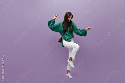 Long-haired brunette woman in green knitted sweater dances on purple background. Happy girl in white pants moves on isolated.