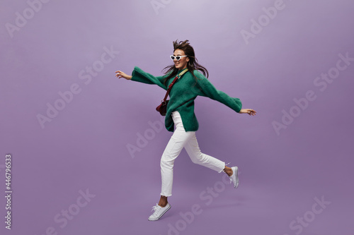 Pretty tanned woman in green sweater and white pants moves on isolated. Charming young girl in sunglasses walks and jumps on purple background.