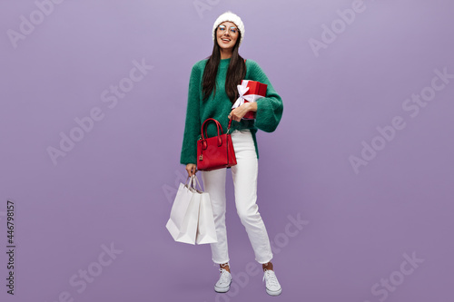 Happy young woman in woolen oversized green sweater, knitted hat, white pants and eyeglasses holds red gift box and shopping bags on purple background.