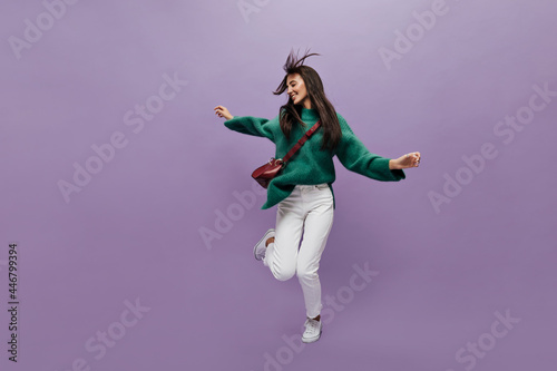 Cheerful woman in white pants and green sweater dances in good mood on purple background. Cheerful girl in stylish outfit moves and jumps on isolated.