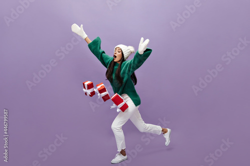 Young woman drops red gift boxes on purple background. Tanned charming girl in green sweater and white pants moves on isolated.