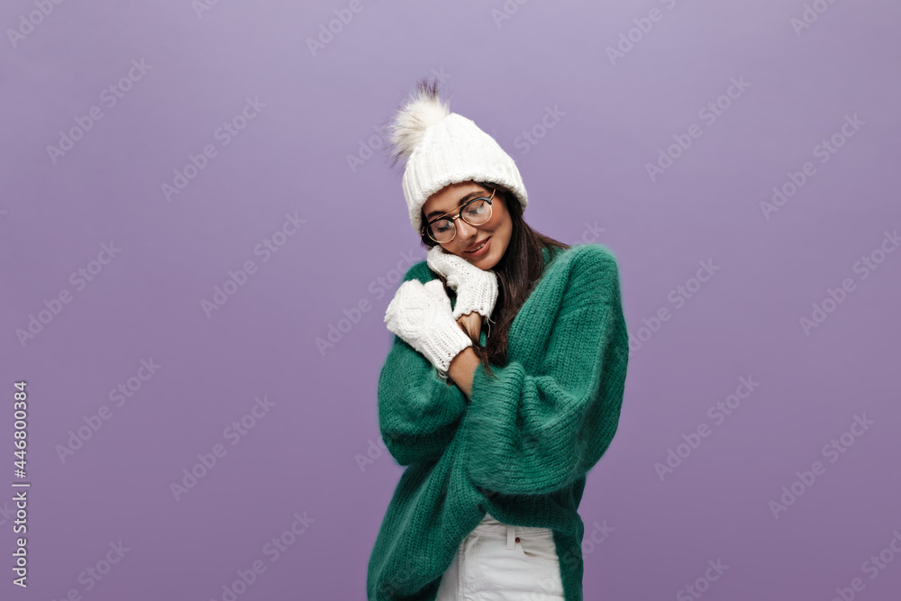 Portrait of shy young brunette woman in white knitted hat and gloves. Pretty long-haired girl in eyeglasses and green sweater poses on purple background.