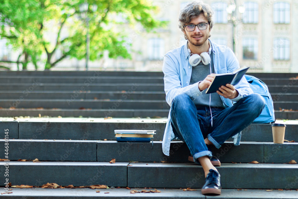 Fototapeta premium Happy university student is reading book while sitting on steps in campus. Caucasian young man is preparing for lessons at college, copy space area for your text message, content. Back to high school