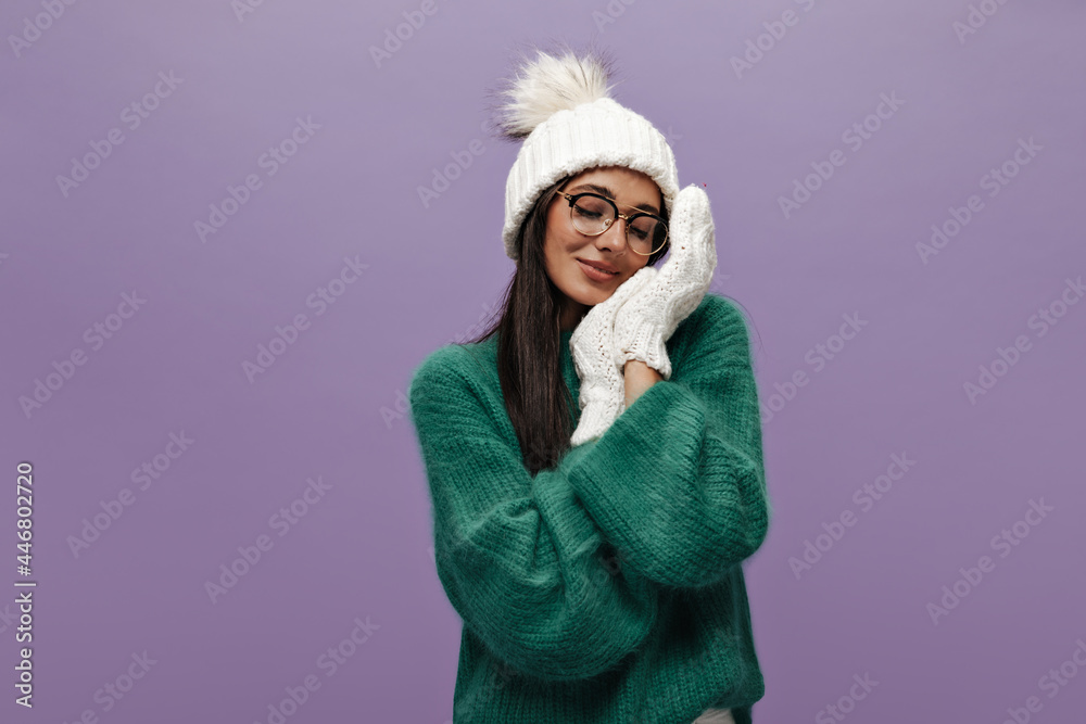 Shy charming girl in warm hat and gloves poses on purple background. Happy lady in green sweater smiles softly on isolated.