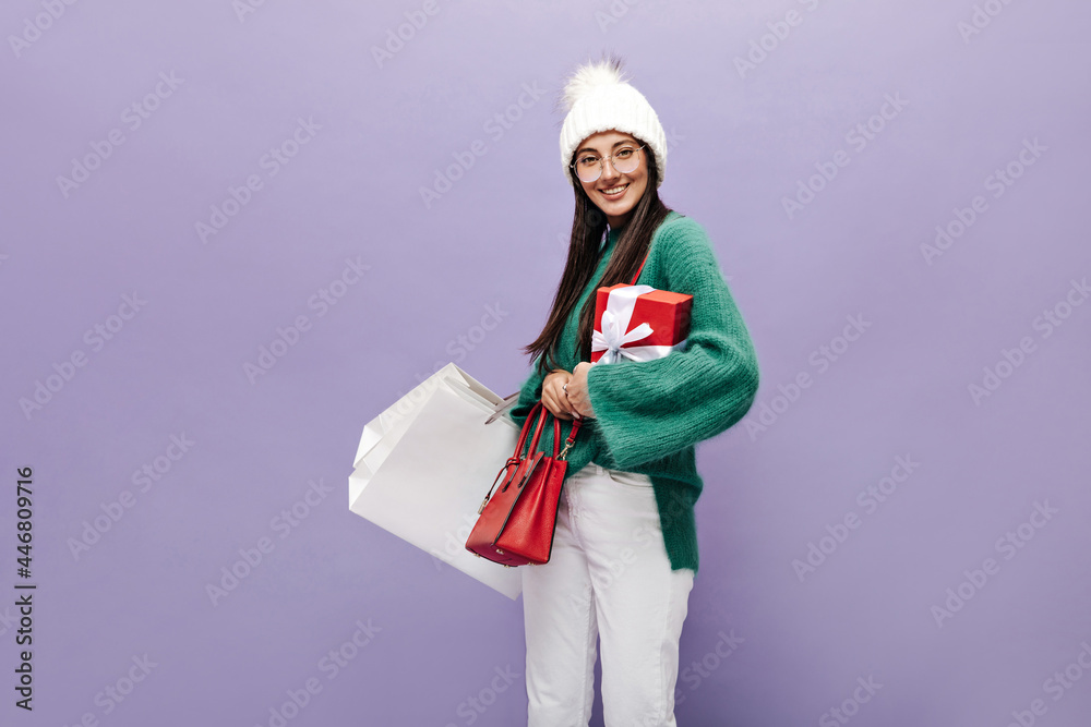 Attractive woman in warm hat, knitted gloves, green woolen sweater and white pants holds gift box, shopping bags and smiles on purple background. Attractive woman in warm hat, knitted gloves, green woolen sweater and white pants holds gift box, shopping bags and smiles on purple background.