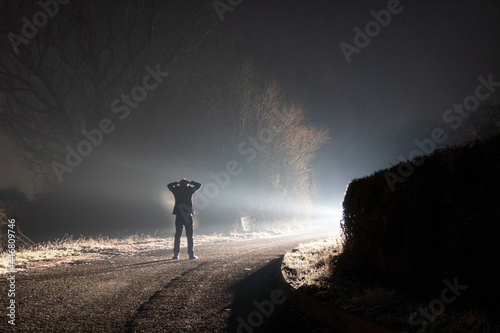 A figure standing on a road surrendering with his hands on his head. On a foggy winters night