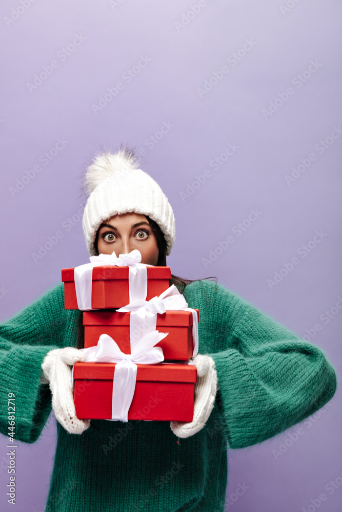 Green-eyed woman in white knitted hat looks into camera. Pretty brunette girl on green woolen sweater holds red gift boxes on purple background. Green-eyed woman in white knitted hat looks into camera. Pretty brunette girl on green woolen sweater holds red gift boxes on purple background.