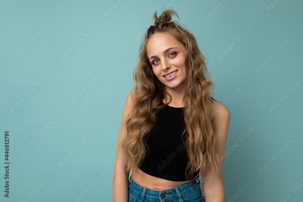 Naklejka premium Young beautiful curly blonde woman with sexy expression, cheerful and happy face wearing trendy black top isolated over blue background with copy space
