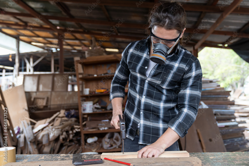 Male carpenter using hammering a nail into wooden board and wearing safety goggles and dust filter mask at the carpentry workshop. Joiner using hammer and nails fixes a wooden plank Male carpenter using hammering a nail into wooden board and wearing safety goggles and dust filter mask at the carpentry workshop. Joiner using hammer and nails fixes a wooden plank