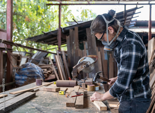 Male carpenter using hammering a nail into wooden board and wearing safety goggles and dust filter mask at the carpentry workshop. Joiner using hammer and nails fixes a wooden plank