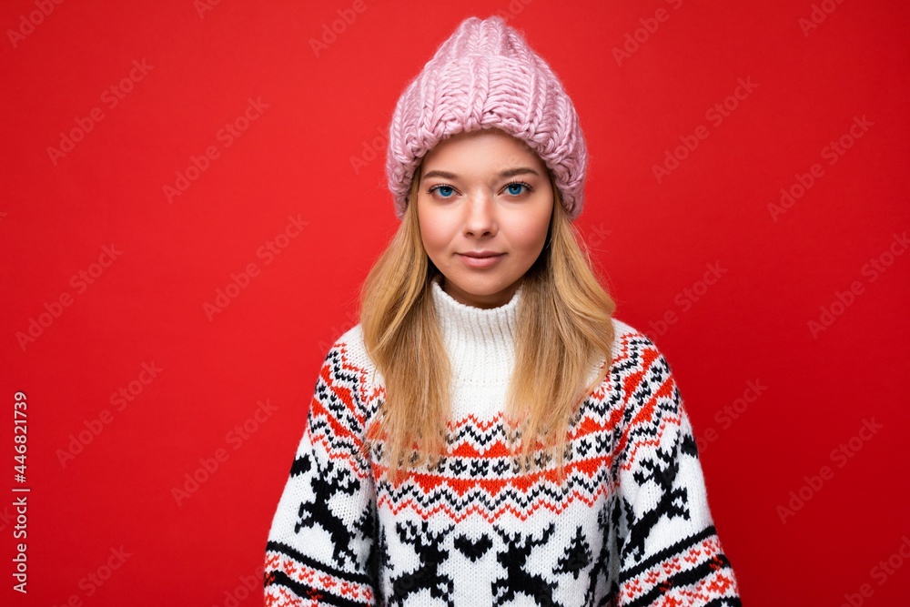 Photo of beautiful calm serious young blonde woman isolated over red background wall wearing winter sweater and pink hat looking at camera Photo of beautiful calm serious young blonde woman isolated over red background wall wearing winter sweater and pink hat looking at camera