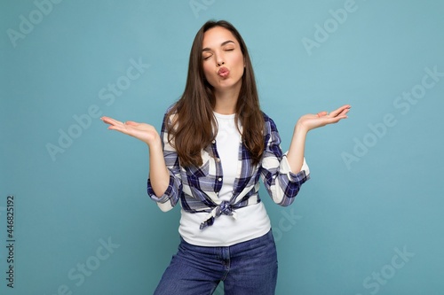 Young positive happy beautiful winsom brunette woman with sincere emotions wearing check shirt poising isolated over blue background with copy space showing two hands and giving kiss