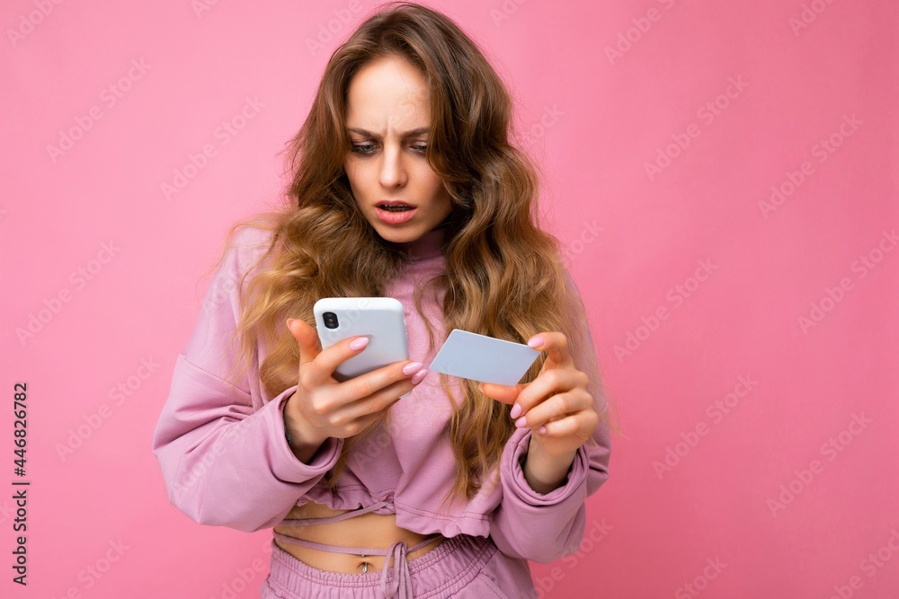Photo of attractive concentrated sad young blonde curly woman wearing pink clothes isolated over pink background using mobile phone making payment using credit card looking at smartphone screen and