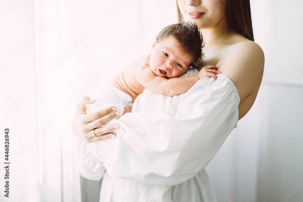 Newborn infant baby girl in a tender embrace of charming mother at the window. Brunette mother holding a small child in the window. Newborn infant baby girl in a tender embrace of charming mother at the window. Brunette mother holding a small child in the window.