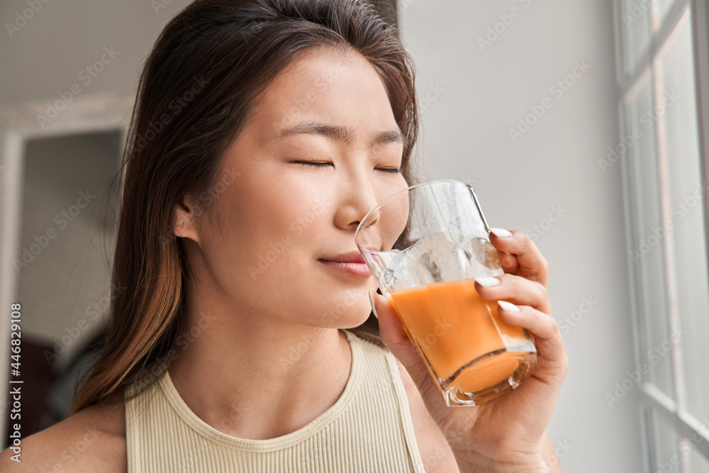 Woman drinking juice while standing near the window at home and closed her eyes Woman drinking juice while standing near the window at home and closed her eyes